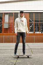 Load image into Gallery viewer, A model stands on a skateboard with a warehouse behind him wearing the Bear Hoodie.
