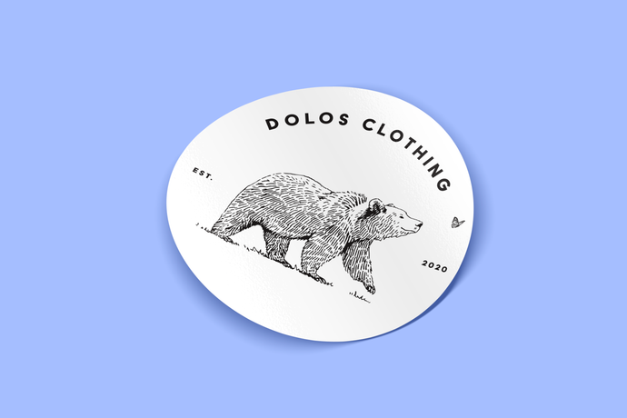 View from an angle - A white sticker with a black bear happily following a butterfly. The sticker says Dolos Clothing on top, and Est. 2020 on the sides
