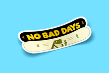 Load image into Gallery viewer, View from an angle - Two snowboards overlapping one another (one white and one tan/yellow). The top board says &quot;no bad days&quot; in yellow writting, with NBD and Dolos on the ends. The tan board has an image of a frog&#39;s head and shoulders with suspenders. NBD and Dolos are written on the sides.
